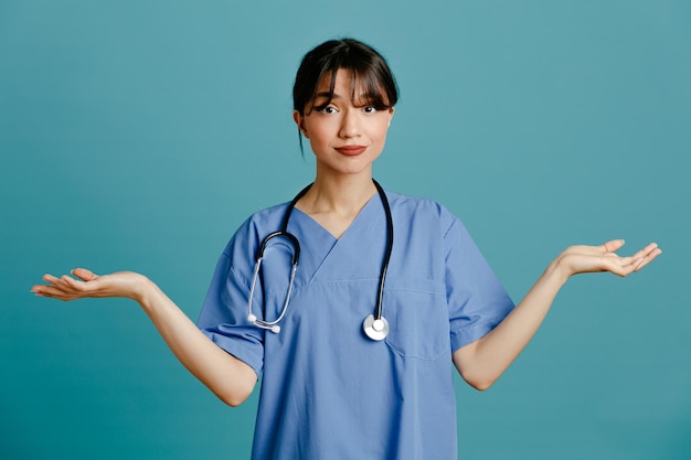 Mani sparse confuse giovane dottoressa che indossa uno stetoscopio fitth uniforme isolato su sfondo blu