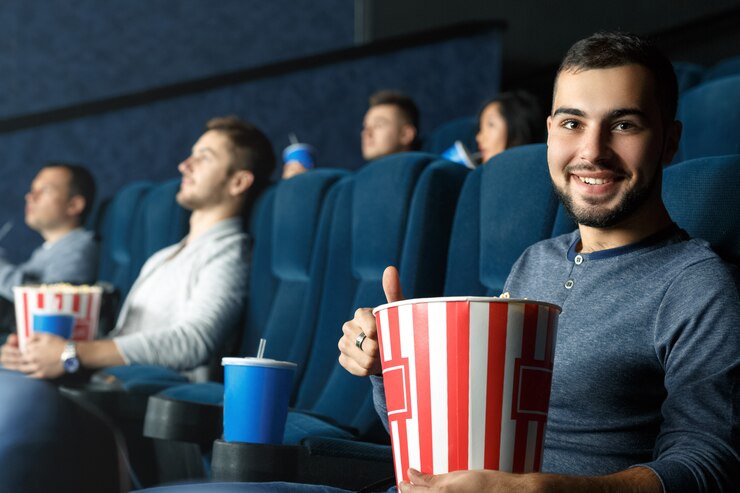 Mania del film. Giovane uomo barbuto bello che mostra i pollici su che si siedono nel cinema che tiene il suo popcorn
