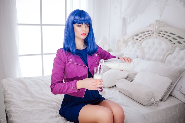 Maniaco della moda. Glamour ragazza sintetica, finta bambola con sguardo vuoto e capelli blu sta versando un latte nel bicchiere mentre è seduta sul letto.