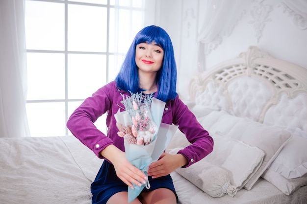Maniaco della moda. Ragazza sintetica sorridente glamour, bambola finta con i capelli blu tiene in mano il mazzo di fiori mentre è seduta sul letto bianco.