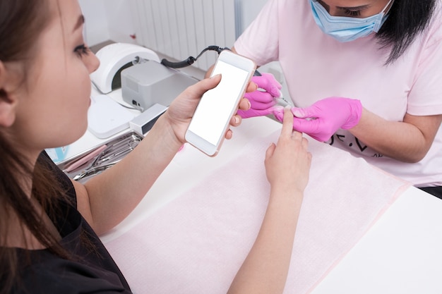 Manicure hardware. Smartphone nelle mani del cliente. Maestro di manicure che fa una procedura di bellezza, ragazza che chiacchiera al telefono