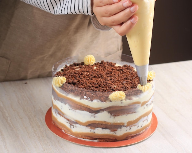 Mano asiatica femminile che decora il tiramisù con il mascarpone di vortice usando la sac à poche per fare la torta intera di tiramisù