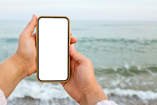 Mano che mostra uno smart phone vuoto sulla spiaggia con il mare sullo sfondo. Schermo bianco mock up