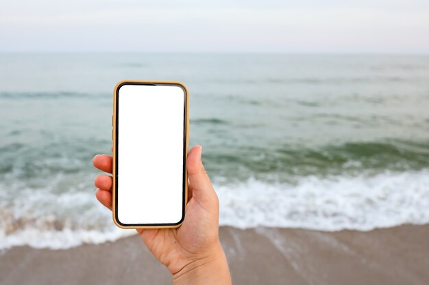 Mano che mostra uno smart phone vuoto sulla spiaggia con il mare sullo sfondo. Schermo bianco mock up