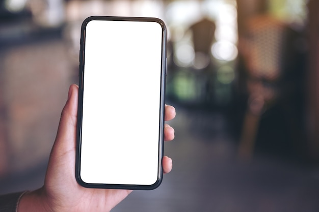 mano che tiene e mostrando il telefono cellulare nero con schermo bianco vuoto nella moderna caffetteria