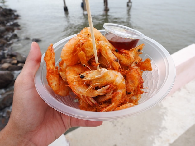 Mano che tiene gamberi fritti croccanti in una ciotola di plastica trasparente, snack tailandese di cibo di strada