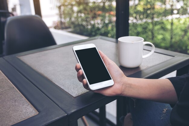 mano che tiene il cellulare bianco con schermo desktop nero bianco con tazza di caffè sul tavolo