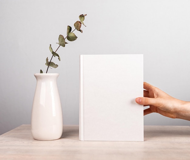 Mano che tiene il libro mockup vicino al vaso con fiori al tavolo di legno Interni belli e teneri di amante della letteratura in stile minimalista