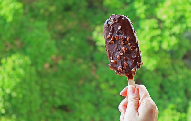 Mano che tiene una barra di gelato immersa nel cioccolato contro il fogliame verde sfocato