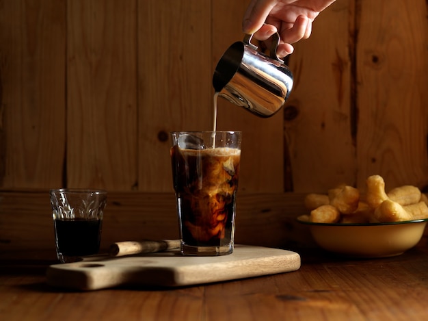 Mano che versa il latte nel caffè ghiacciato sulla tavola di legno con bastoncini di pasta fritta