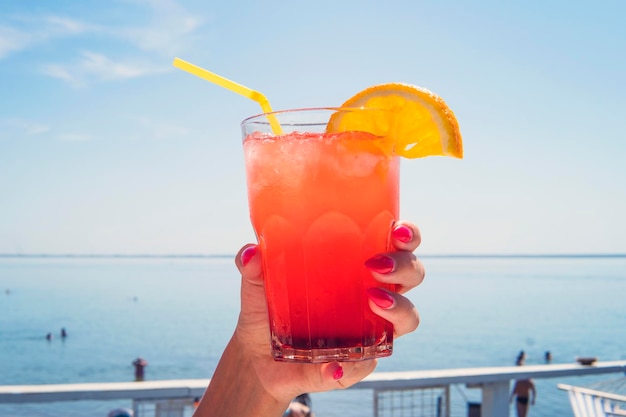 Mano con bicchiere da cocktail arancione sulla baia esotica della laguna turchese Stile di vita concetto di vacanza estiva ragazza che tiene un bicchiere di succo sullo sfondo di fare il bagno in acqua turisti sulla spiaggia in estate