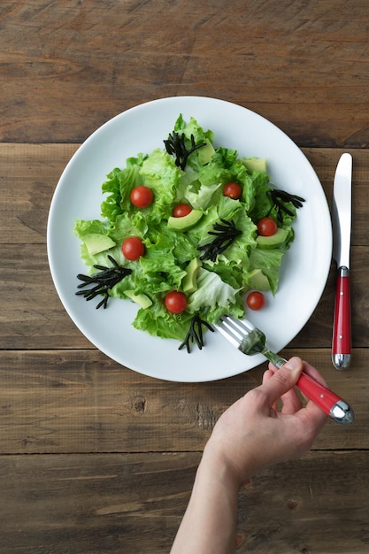 Mano con forchetta che mangia insalata assortita su piatto bianco su sfondo di legno Copia spazio Foto verticale