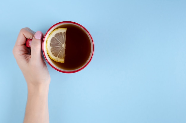 Mano con la composizione rossa nella fetta della tazza e del limone di tè su fondo blu.