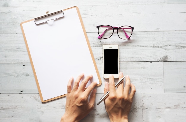 Mano con penna su appunti e occhiali e utilizzando il display nero del cellulare sulla vista del piano del tavolo in legno