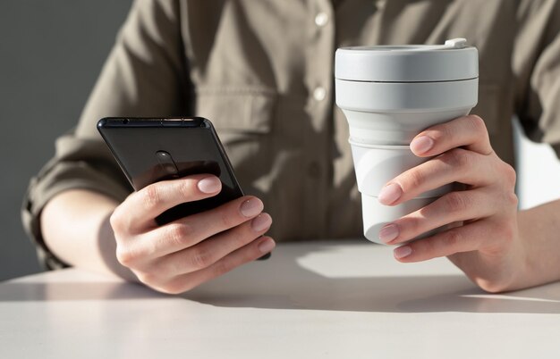 Mano con tazza ecologica e telefono cellulare al tavolo da vicino