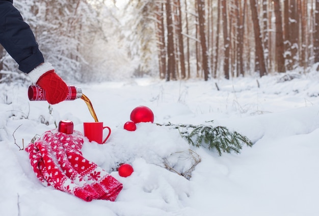 Mano con tè in thermos rosso nella foresta invernale