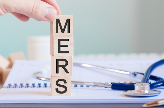 Mano d'uomo che tiene un cubo con la lettera M dalla parola MERS scritta su cubi di legno vicino a uno stetoscopio su uno sfondo di carta, concetto medico