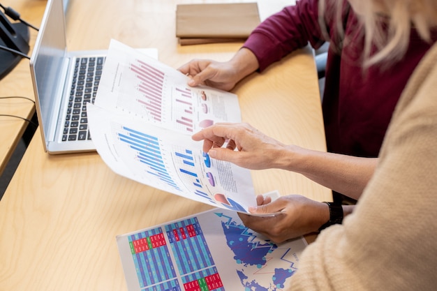 Mano del mediatore femminile contemporaneo che indica uno dei documenti finanziari detenuti dal collega mentre discuteva il grafico e il grafico dallo scrittorio