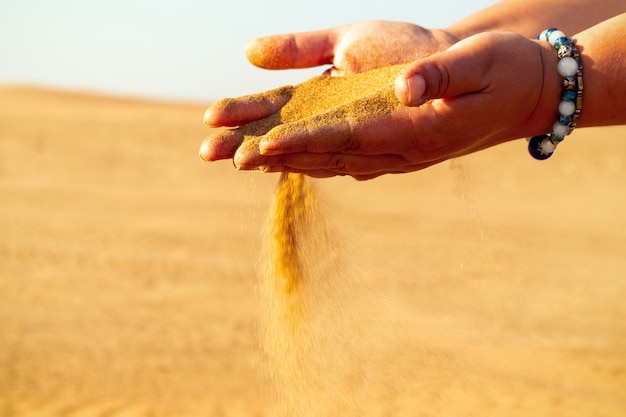 Mano del primo piano che rilascia sabbia cadente. Sabbia che scorre tra le mani contro il deserto dorato. Vacanze estive.