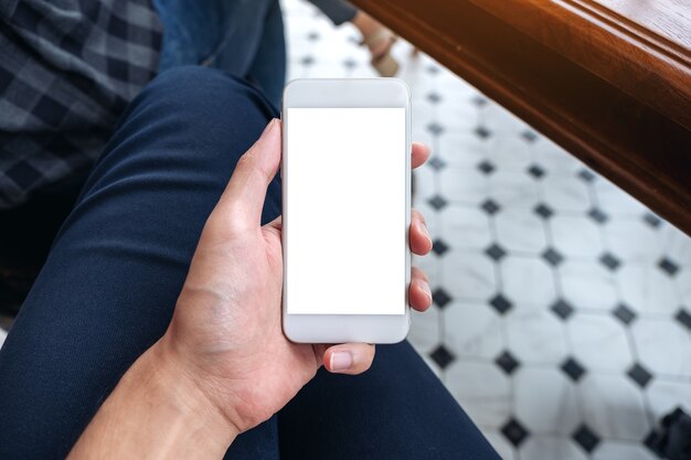 mano dell'uomo che tiene il telefono cellulare bianco con schermo del desktop vuoto sulla coscia nella caffetteria