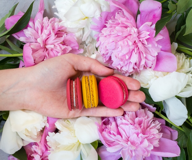 Mano della donna che tiene amaretti sul tavolo con peonie rosa, vista dall'alto, piatto laici