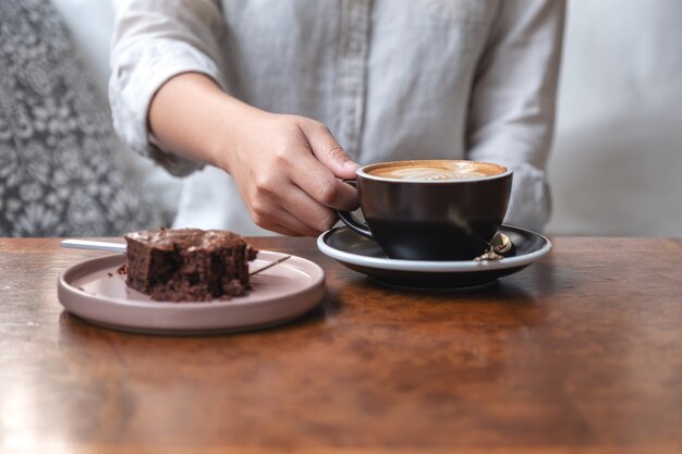 mano della donna che tiene e beve caffè latte caldo con torta brownie sul tavolo