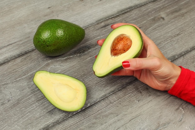 Mano della donna che tiene l'avocado tagliato a metà, con l'altro intero su un tavolo di legno grigio in bacgkground.