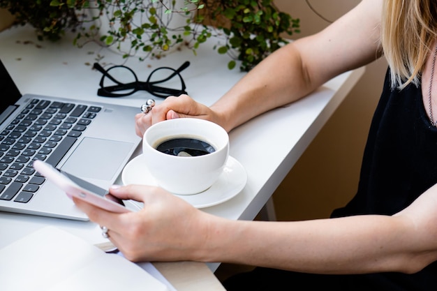 Mano della donna che tiene tazza di caffè e smartphone in ufficio accogliente con computer portatile e piante verdi