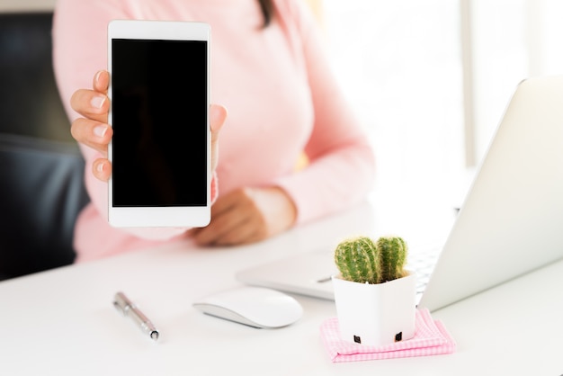 Mano della donna che tiene telefono cellulare bianco su un tavolo con un computer portatile in ufficio