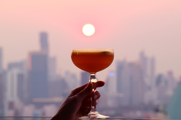 Mano della donna che tiene un bicchiere da cocktail contro il sole al tramonto con grattacieli sfocati sullo sfondo