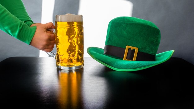 Mano della donna che tiene un bicchiere di birra vicino al cappello di giorno di San Patrizio di un leprechaun sul nero