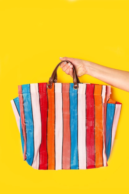 Mano della donna che tiene una borsa della spesa di plastica a strisce colorate su uno sfondo giallo