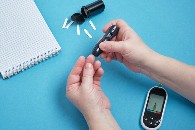 Mano della donna che utilizza una lancetta sul dito per controllare lo zucchero nel sangue utilizzando un glucometro, diabete, glicemia, concetto di assistenza sanitaria. sfondo blu copia spazio vista dall'alto