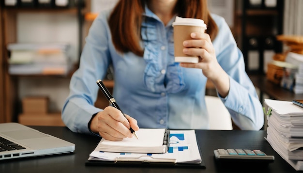Mano della donna di affari che scrive sul blocco note con il nuovo diagramma di strategia del computer moderno e che tiene la tazza di caffè come luce del mattino di concetto