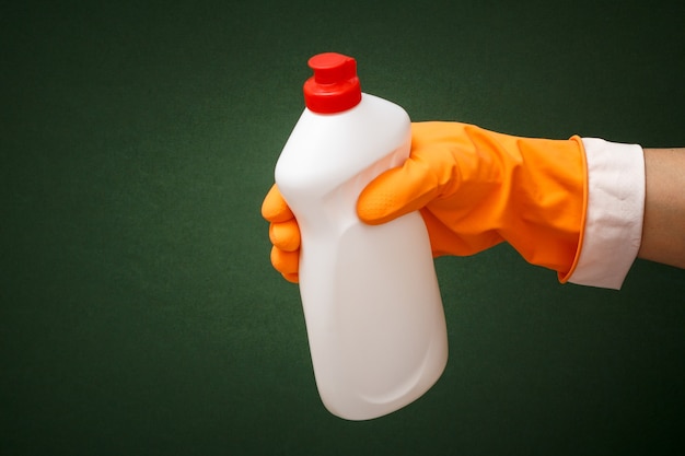 Mano della donna in guanto protettivo arancione con bottiglia di detersivo per piatti su sfondo verde. Concetto di lavaggio e pulizia.