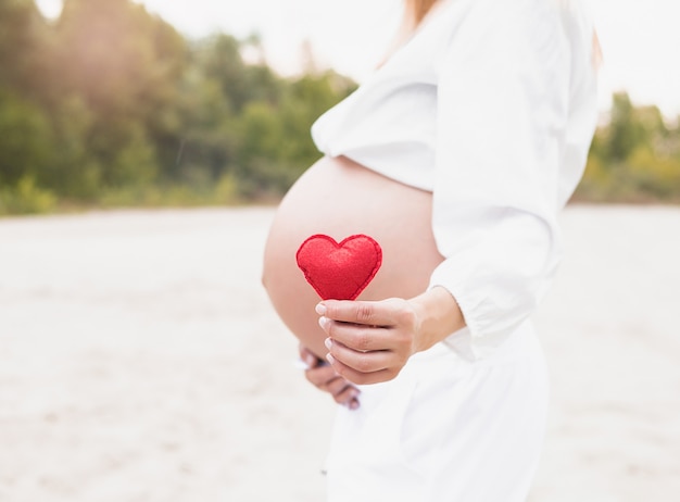 Mano della giovane donna che tiene forma luminosa rossa del cuore sulla pancia nuda