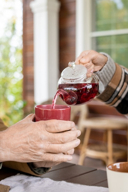 Mano della moglie anziana con teiera che versa tè alle erbe nero nella tazza di ceramica tenuta da suo marito sul tavolo servito per colazione all'aperto