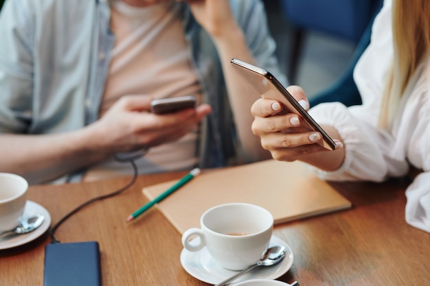 Mano di giovane donna mobile con smartphone scorrendo i dati online o contatti sul tavolo mentre sorseggia una tazza di tè