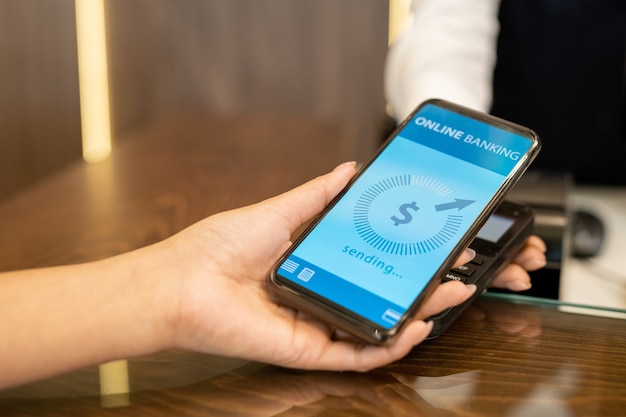 Mano di una giovane viaggiatrice che tiene in mano uno smartphone con una pagina bancaria online sullo schermo sopra la macchina di pagamento in piedi sul bancone della reception