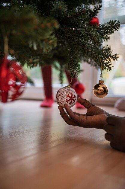 Mano di una ragazza di colore che tiene una pallina di festa rossa e bianca appesa a un albero di natale.