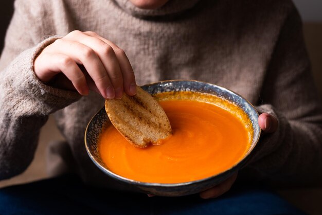 Mano femminile che immerge un pezzo di pane in un piatto con zuppa di crema di zucca, cibo vegetale sano.