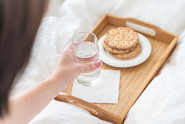 Mano femminile che tiene bicchiere d'acqua sopra il vassoio con i cracker sul letto