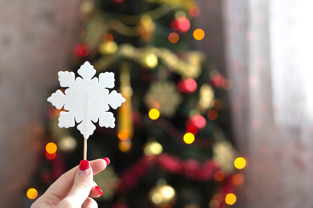 Mano femminile che tiene il fiocco di neve di carta bianca sullo sfondo dell'albero di natale con luci