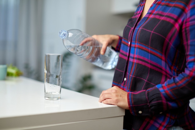 Mano femminile che tiene la bottiglia dell'acqua potabile e versa l'acqua nel bicchiere sul tavolo sullo sfondo della cucina sfocata.