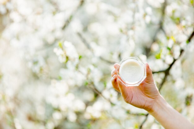 Mano femminile che tiene la crema per la cura della pelle vicino all'albero in fiore. Stagione primaverile