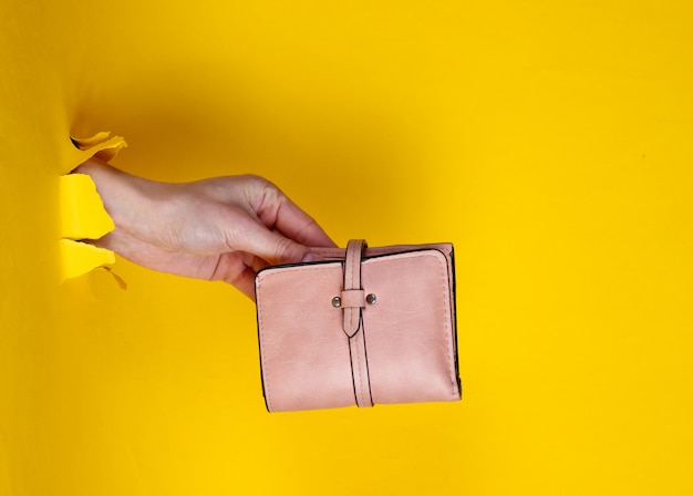 Mano femminile che tiene portafoglio rosa attraverso carta gialla lacerata. Concetto di moda creativa minimalista