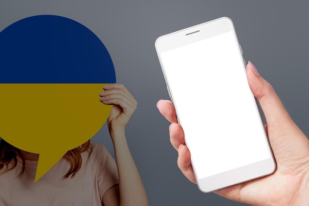 Mano femminile che tiene un telefono cellulare con uno schermo bianco vuoto e una donna che tiene un poster di carta con l'immagine della bandiera ucraina isolata su uno sfondo grigio
