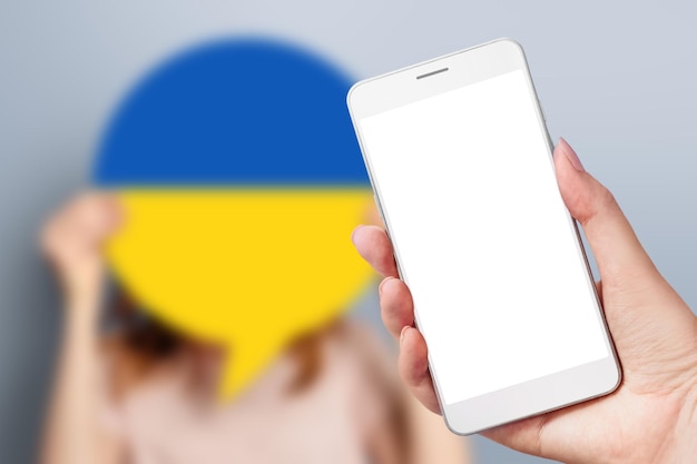 Mano femminile che tiene un telefono cellulare con uno schermo bianco vuoto e una donna che tiene un poster di carta con l'immagine della bandiera ucraina isolata su uno sfondo grigio