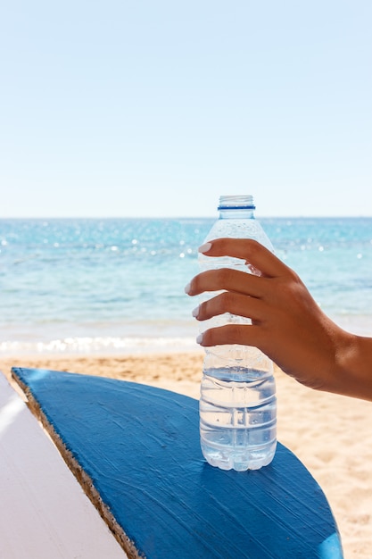 Mano femminile che tiene una bottiglia di acqua sulla spiaggia, fondo del mare