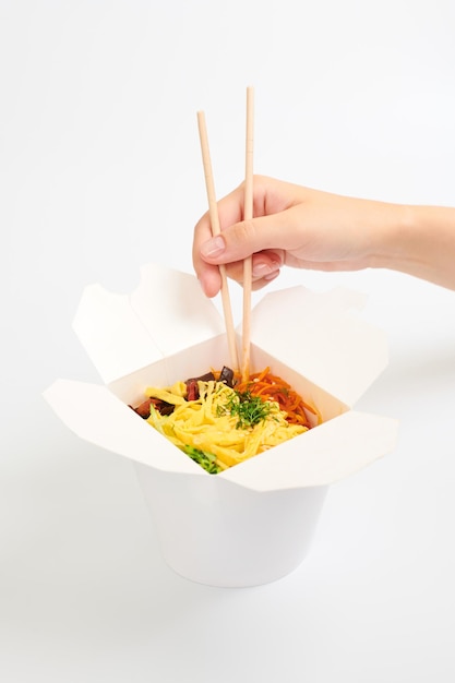 Mano femminile che tiene wok di riso con bacchette di legno, wok cinese asiatico isolato su sfondo bianco. Una scatola da asporto aperta con wok, cocktail di frutti di mare e verdure. Concetto di consegna fast food